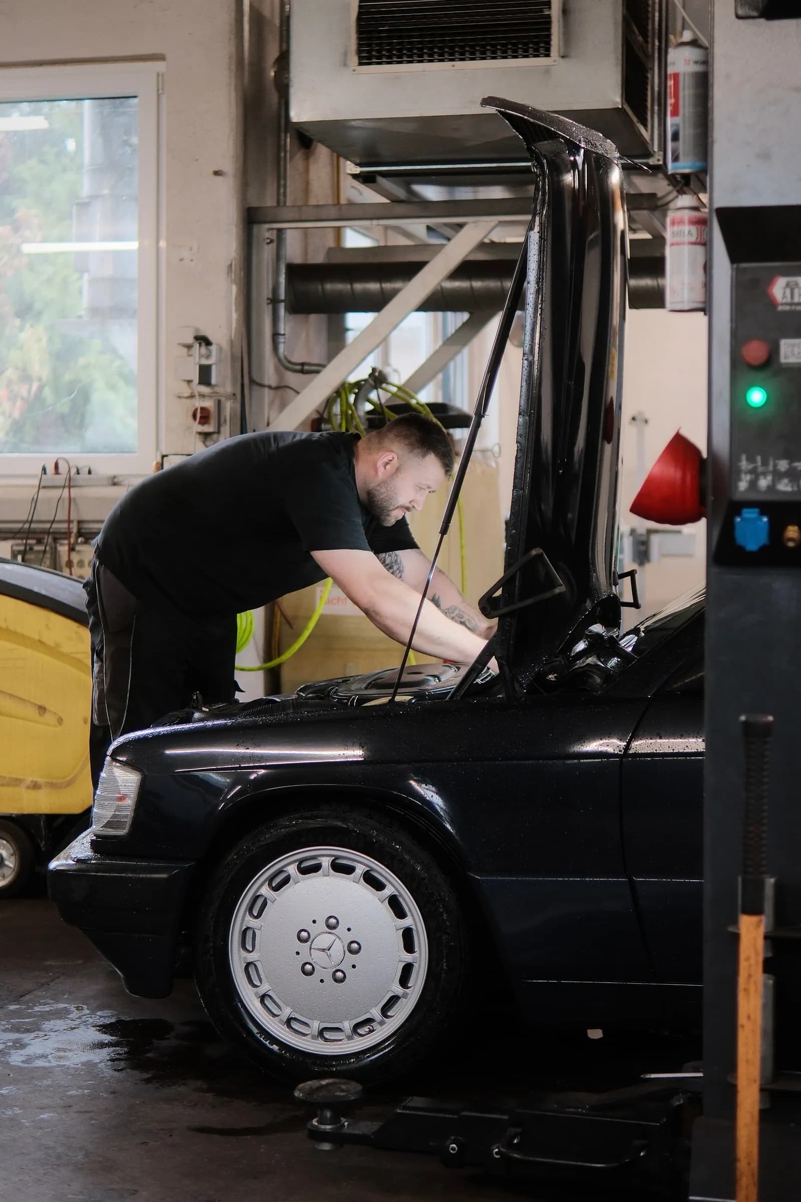 Kfz-Meister Kai Block beugt sich unter der geöffneten Motorhaube über den Motorraum eines schwarzen Mercedes 190