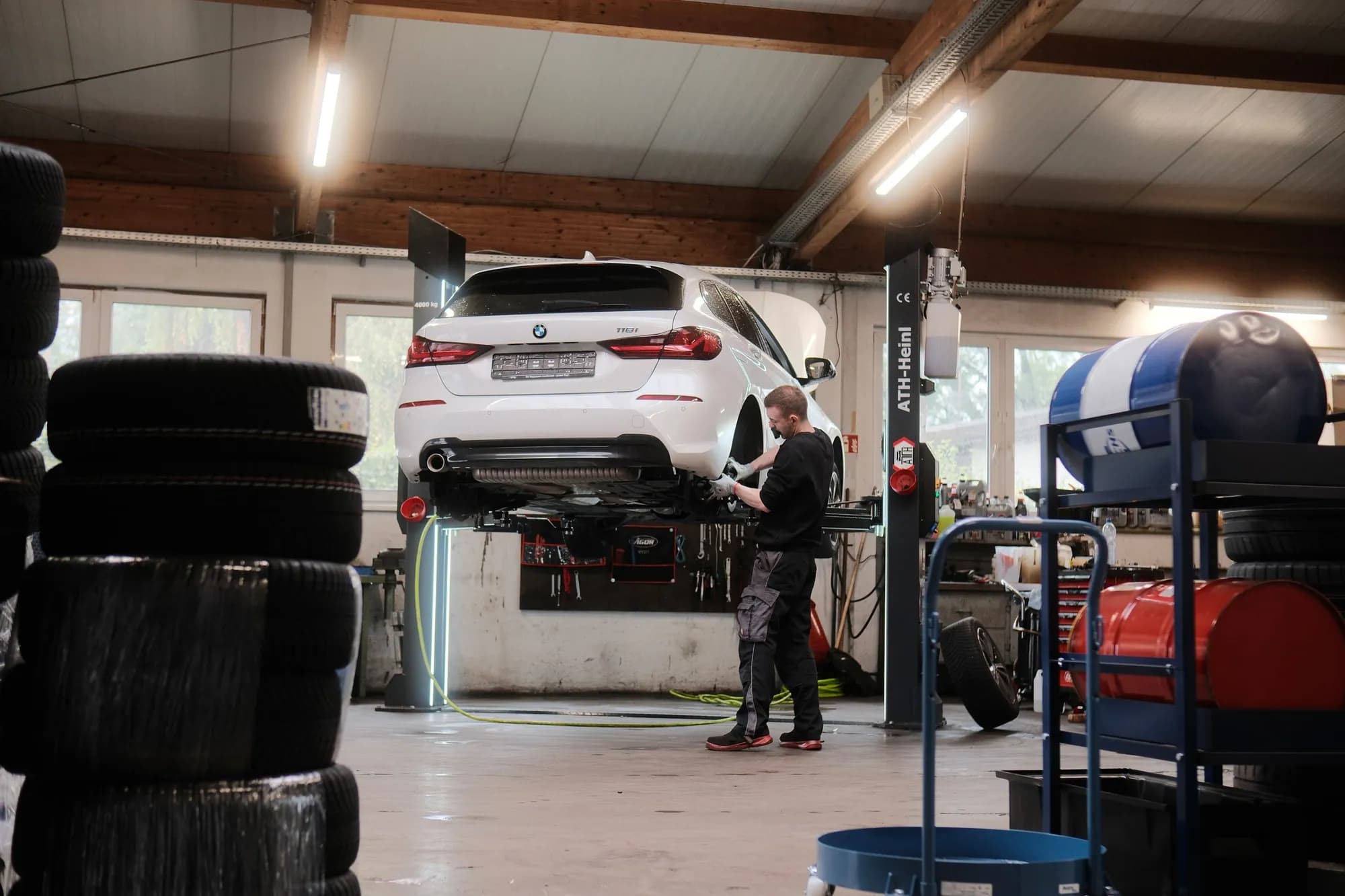 Ein Mechaniker prüft von unten das Fahrwerk eines weißen BMW, der auf der Zwei-Säulen-Hebebühne angehoben ist; im Vordergrund ein Stapel Reifen