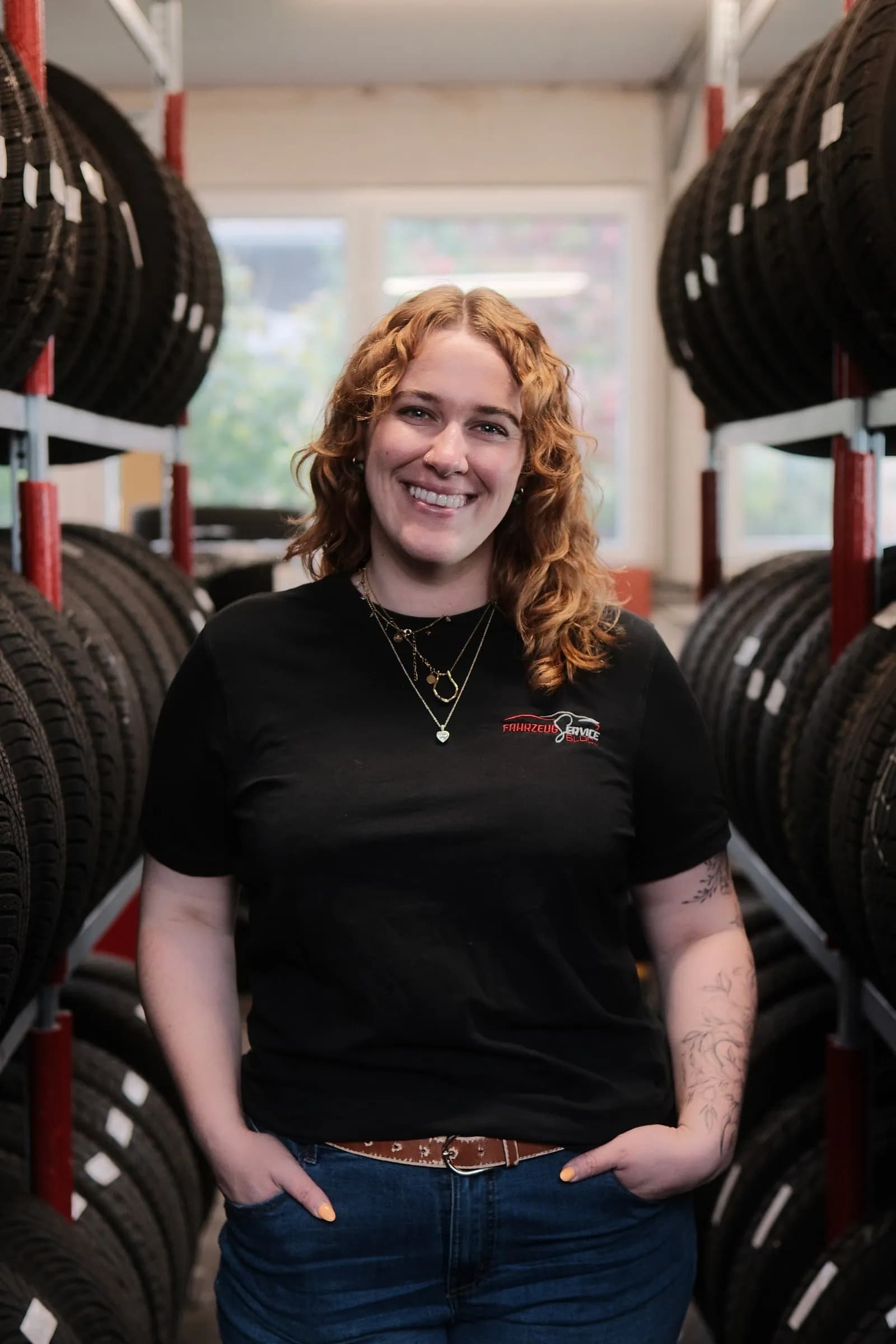 Anna steht lächelnd im Reifenlager zwischen zwei hohen Regalen voller Reifen, im T-Shirt mit dem Logo von Fahrzeugservice Block