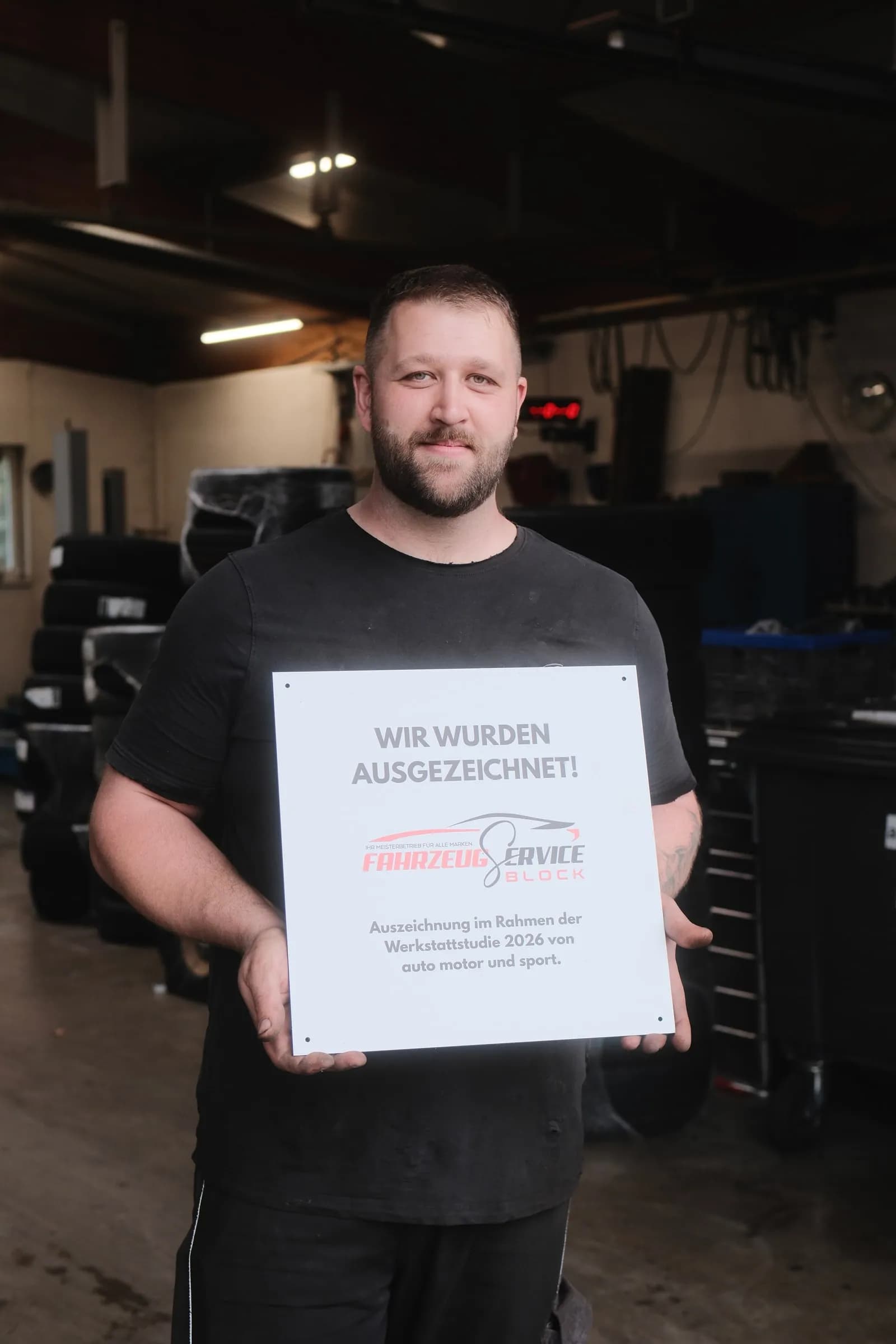 Kai Block steht in der Werkstatthalle und hält ein Schild: „Wir wurden ausgezeichnet!“, darunter das Logo von Fahrzeugservice Block und „Auszeichnung im Rahmen der Werkstattstudie 2026 von auto motor und sport“