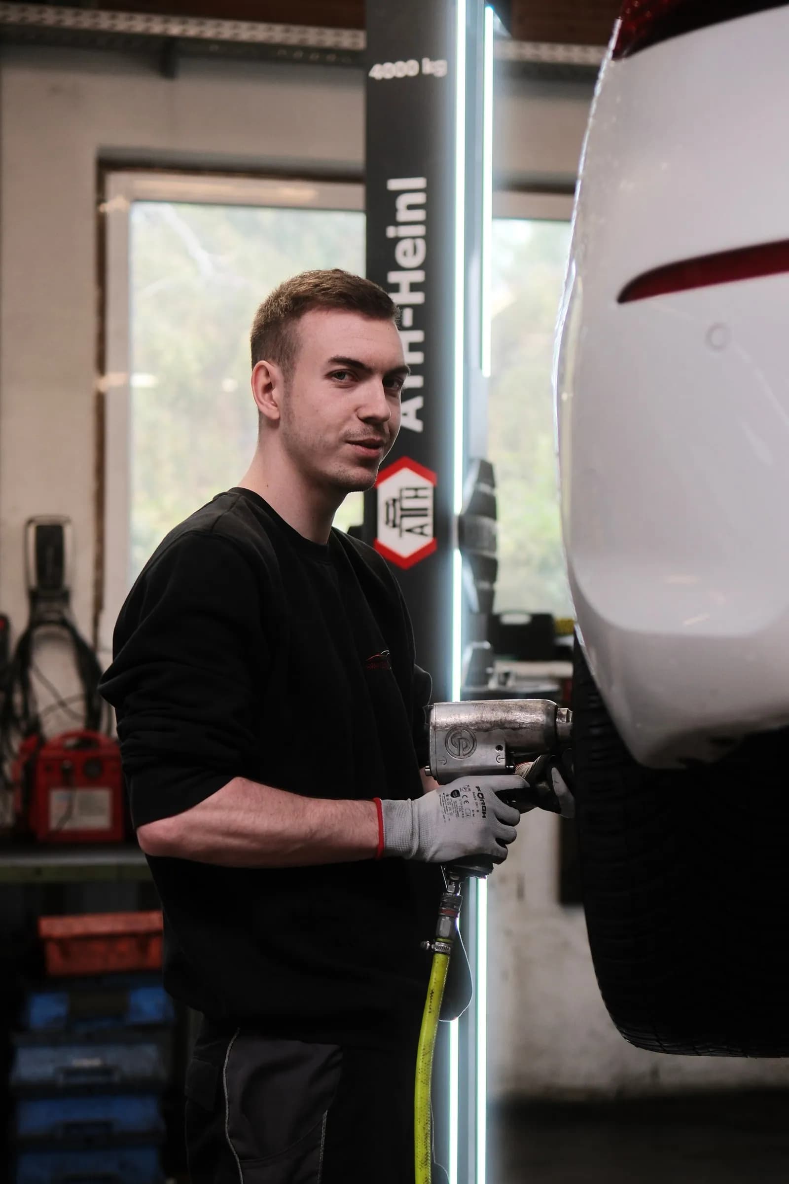Kevin hält mit Arbeitshandschuhen den Schlagschrauber an das Rad eines angehobenen weißen Fahrzeugs und schaut zur Kamera