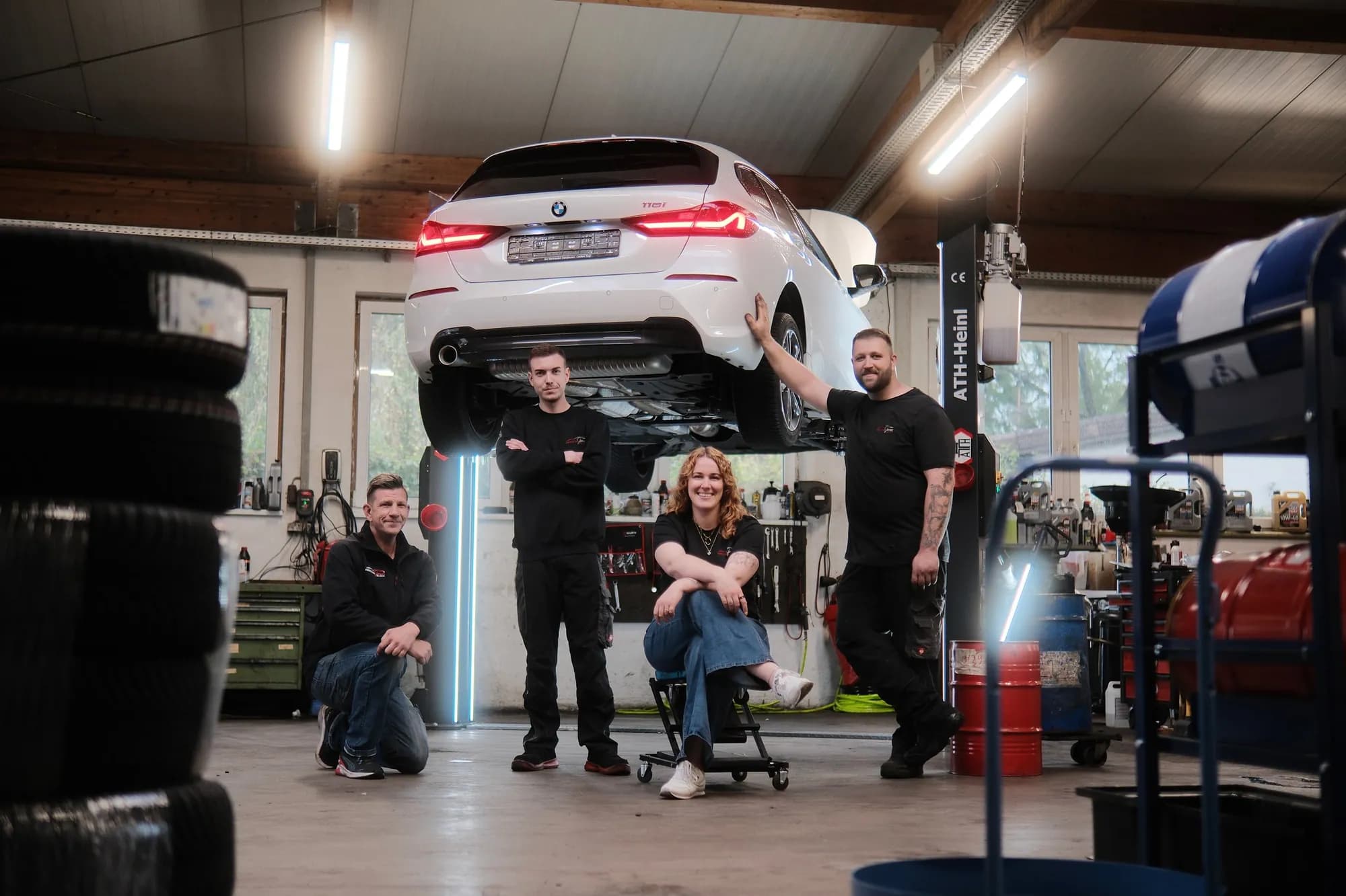 Das Team in der Werkstatthalle unter einem angehobenen weißen BMW: Mikha kniet links, daneben steht Kevin mit verschränkten Armen, Anna sitzt auf einem Rollhocker, Kai stützt sich rechts an das Fahrzeug