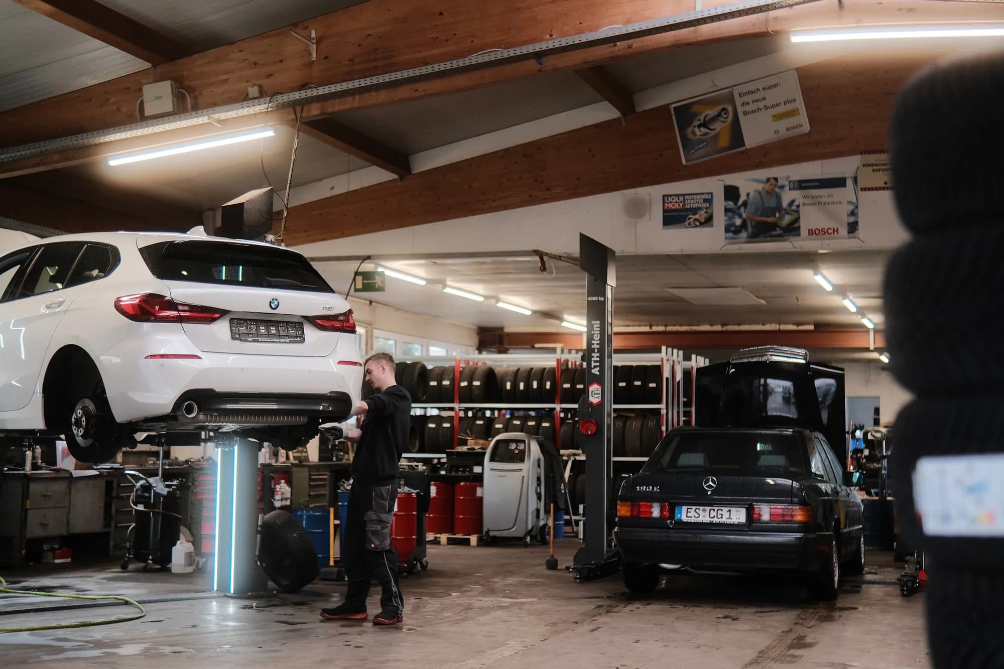 Blick in die Werkstatthalle: ein Mechaniker arbeitet am Rad eines weißen BMW auf der Zwei-Säulen-Hebebühne, dahinter das Reifenregal und Werbeschilder an der Wand, rechts ein schwarzer Mercedes 190
